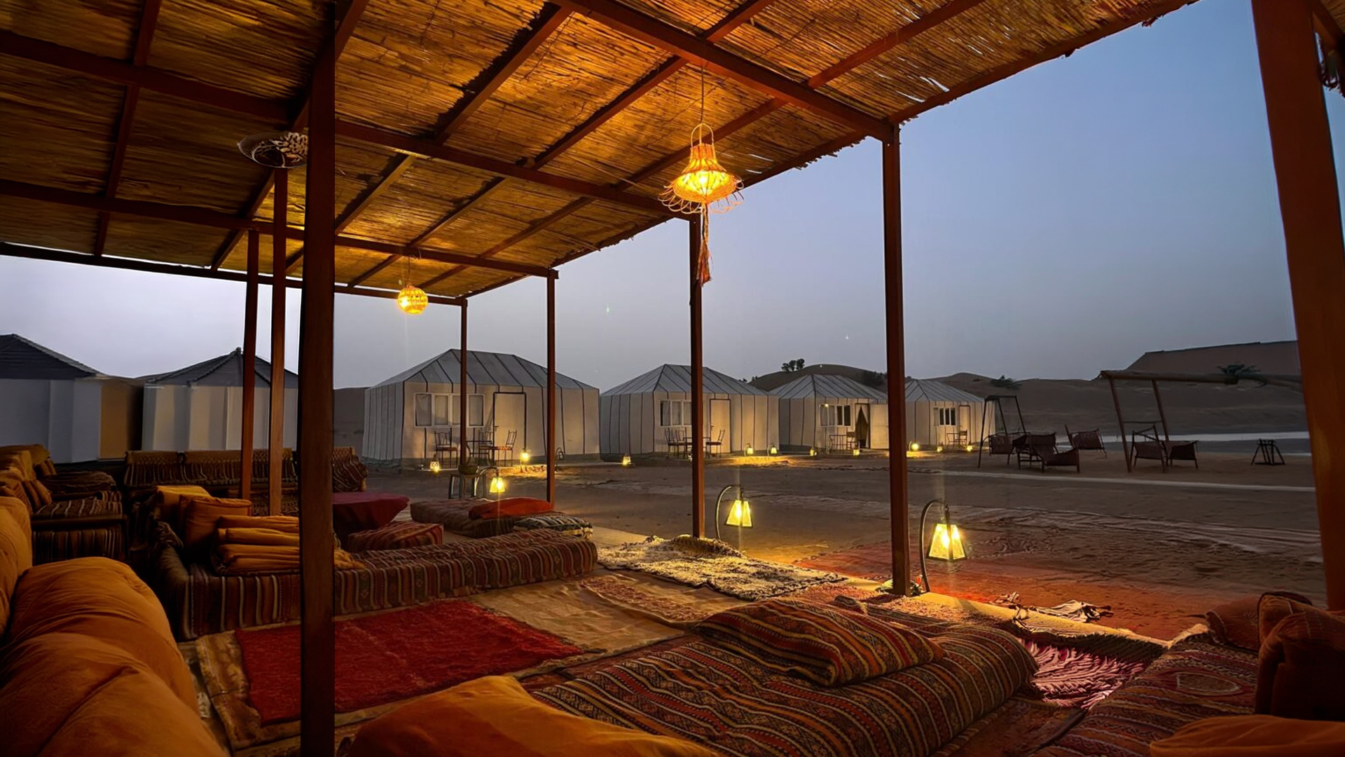 Salon berbère sous la paillote au crépuscule — coussins, tapis, lanternes
